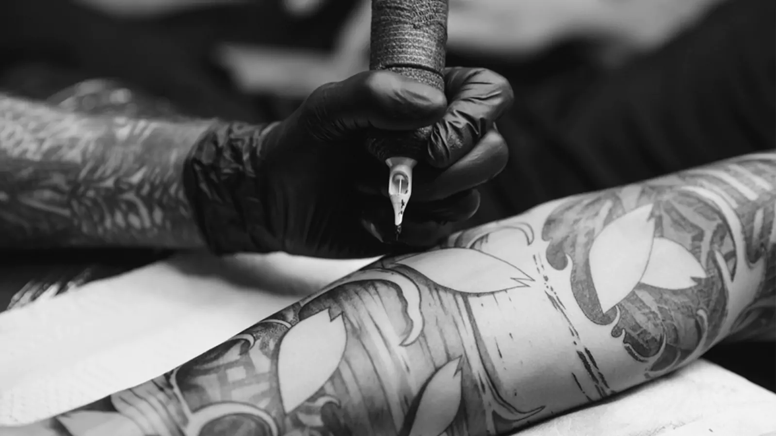 A close-up shot of a tattoo artist's gloved hand as they carefully work on a detailed tattoo on a client's forearm. The tattoo design features bold lines and patterns, with large sections that appear to be in the process of being filled in.